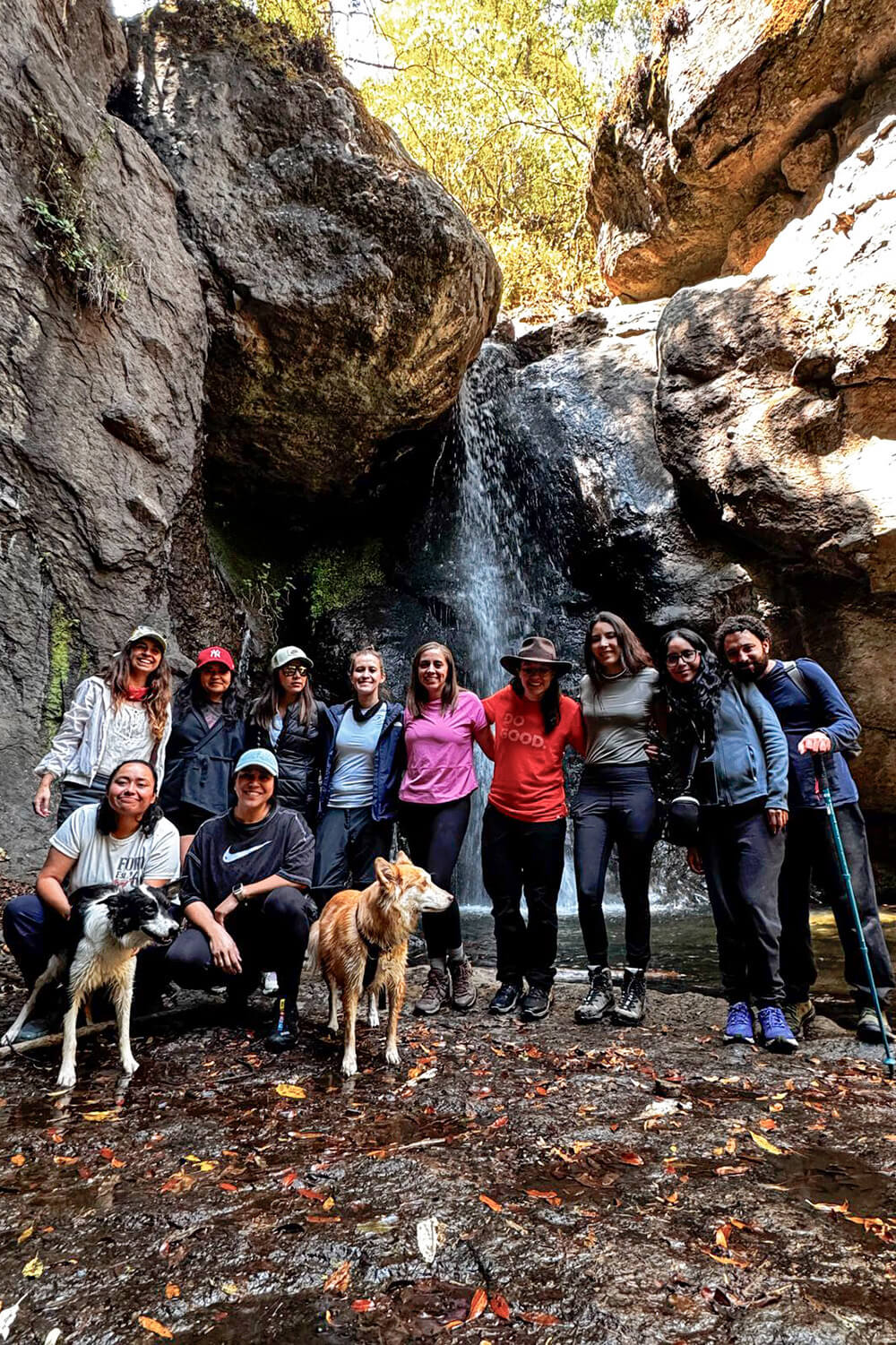 Un grupo de personas posa frente a una cascada, rodeado de terreno rocoso y árboles. Dos perros aparecen en primer plano. El grupo viste ropa informal de calle y algunos sostienen bastones de senderismo. La luz del sol se filtra entre los árboles.